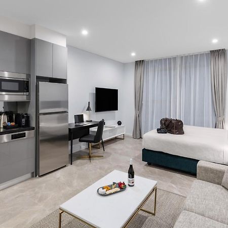 Meriton Suites Sussex Street, Sydney Zewnętrze zdjęcie The photo shows a modern studio apartment. On one side, there is a kitchenette with sleek gray cabinets, a refrigerator, and built-in appliances. In the living area, there is a comfortable gray sofa, a white coffee table with a fruit platter and a bo