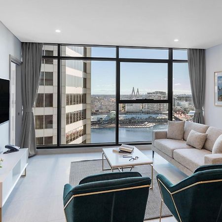 Meriton Suites Sussex Street, Sydney Zewnętrze zdjęcie The photo shows a modern living room with large windows that offer a view of the cityscape and water. The interior features a light color palette with pale blue walls and soft lighting. There is a comfortable gray sofa and two green accent chairs, ar