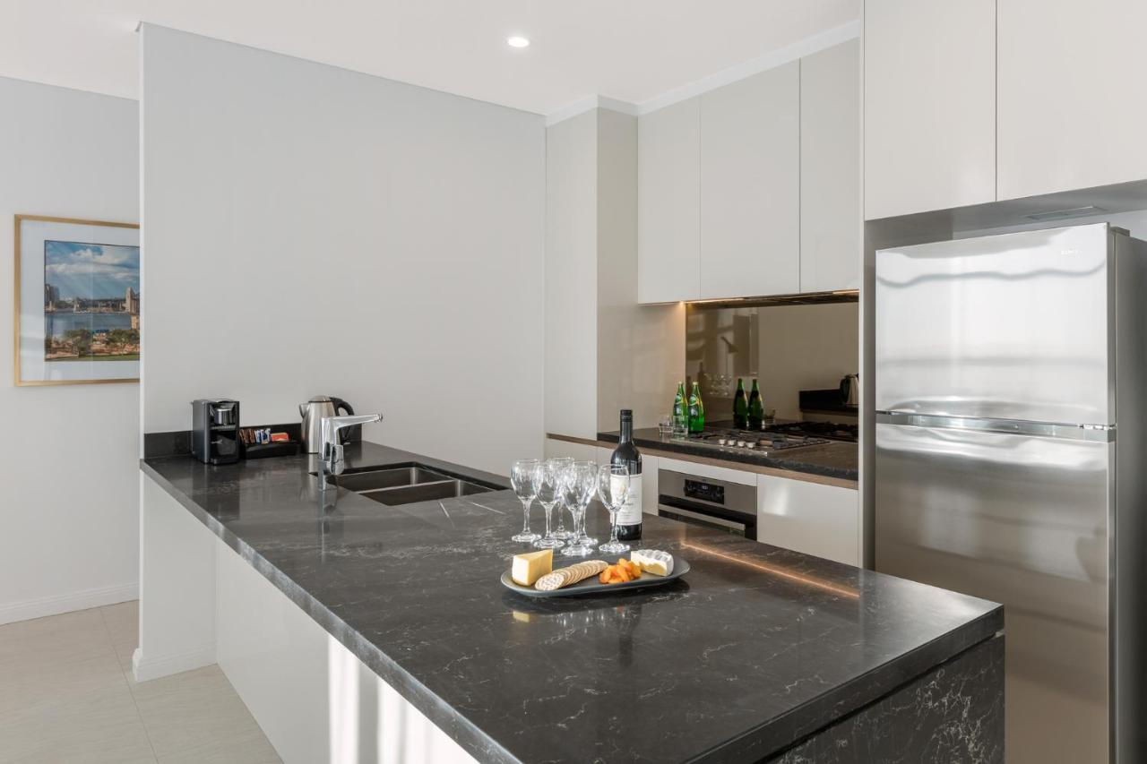 Meriton Suites Sussex Street, Sydney Zewnętrze zdjęcie The photo shows a modern kitchen featuring a sleek design with a dark stone countertop. There is a built-in sink and possibly a cooktop. The kitchen cabinetry is light-colored, creating a contrast with the dark counter. On the countertop, there are w