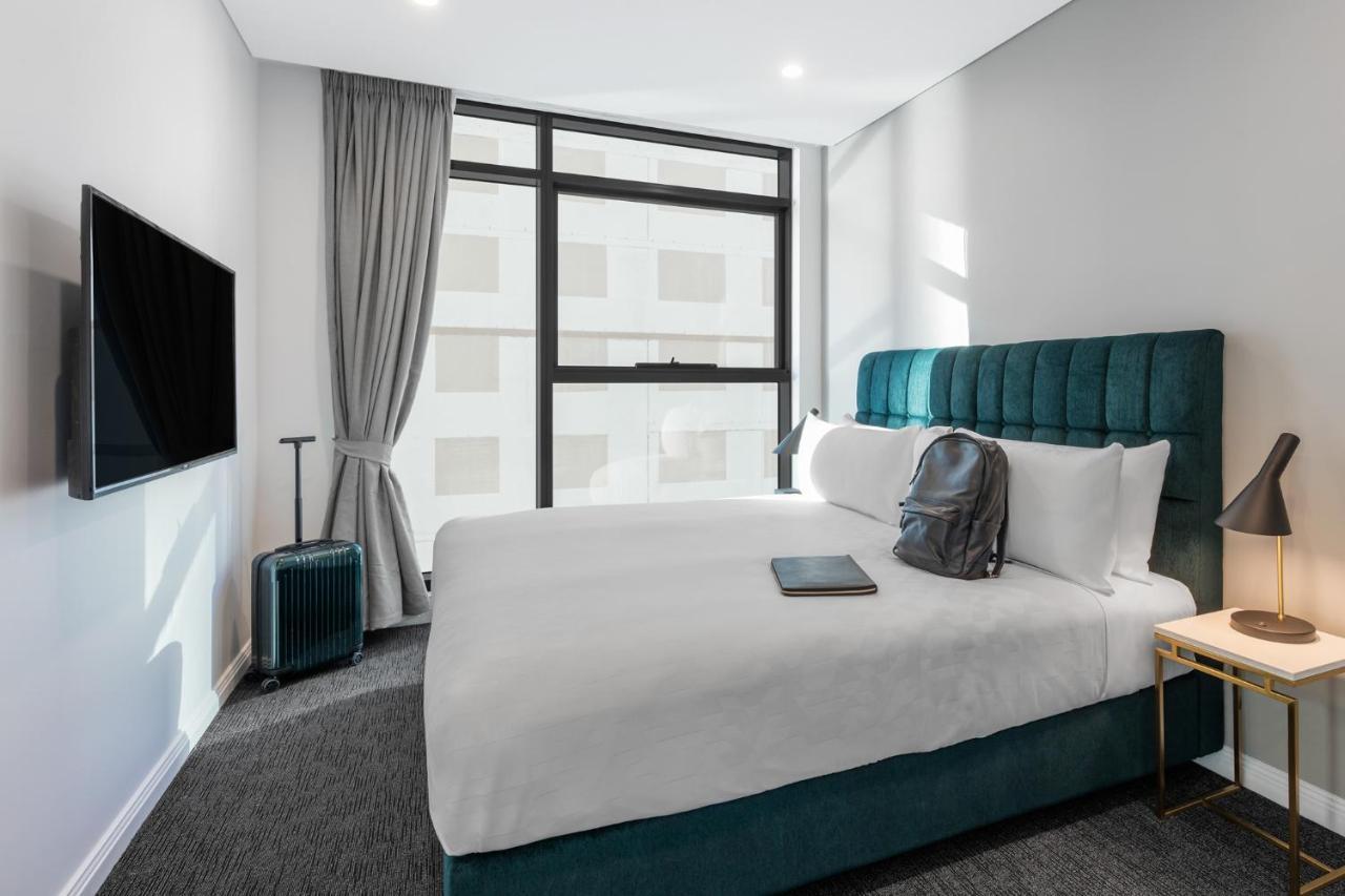 Meriton Suites Sussex Street, Sydney Zewnętrze zdjęcie The photo shows a modern hotel room featuring a comfortable bed with white bedding and a teal-colored headboard. There is a neatly placed black backpack on the bed, along with a small notebook or tablet. A television is mounted on the wall opposite t