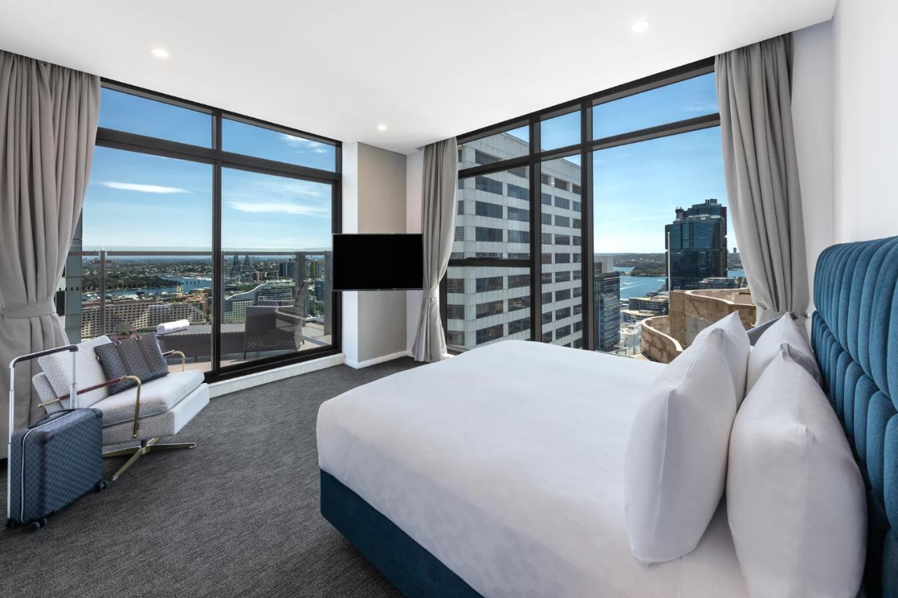 Meriton Suites Sussex Street, Sydney Zewnętrze zdjęcie The photo shows a modern hotel room featuring large windows that provide a panoramic view of the cityscape and waterfront. The room is furnished with a king-size bed dressed in white linens, a comfortable armchair, and a flat-screen TV mounted on the