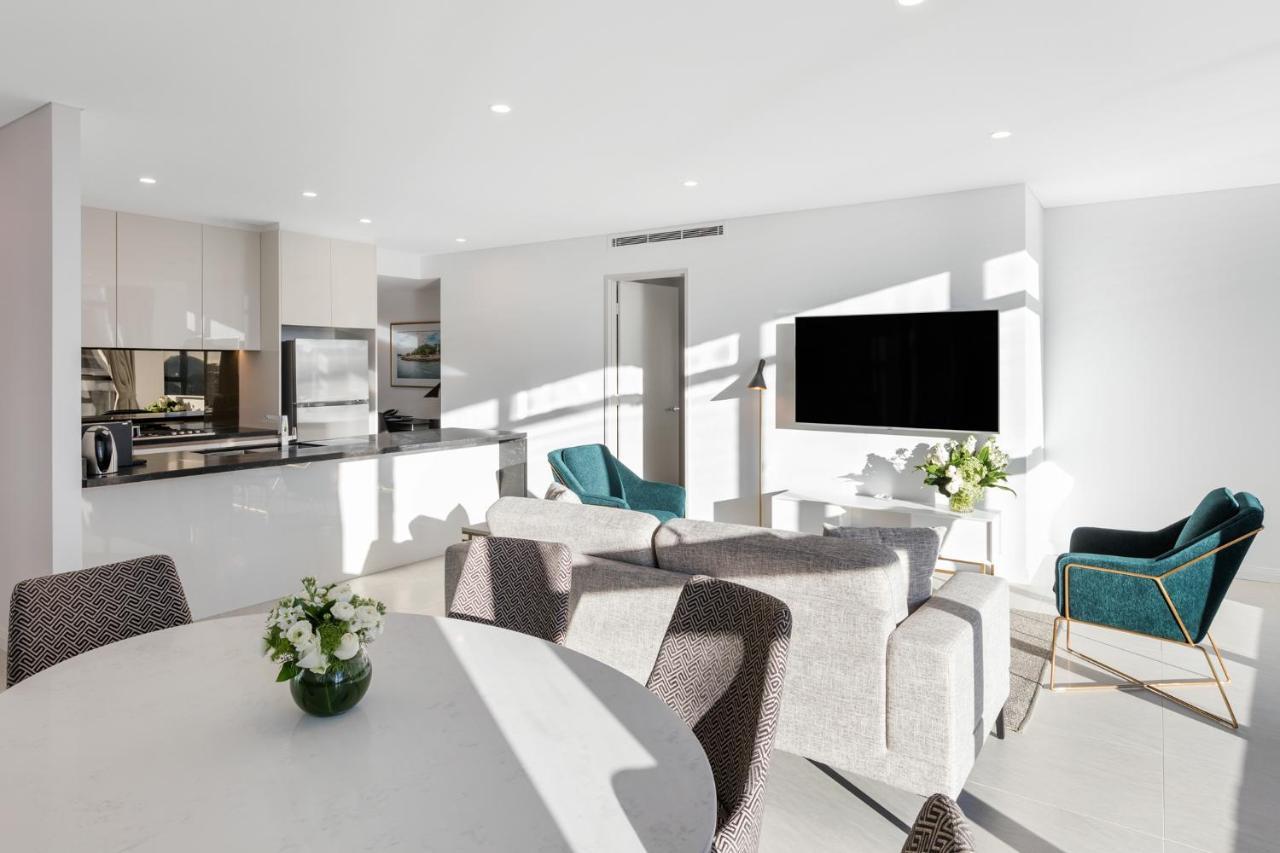 Meriton Suites Sussex Street, Sydney Zewnętrze zdjęcie The photo shows a modern, bright living space that combines a kitchen and a living area. The kitchen features sleek cabinets and stainless steel appliances. In the foreground, there is a round dining table with a vase of flowers on it. Seating option