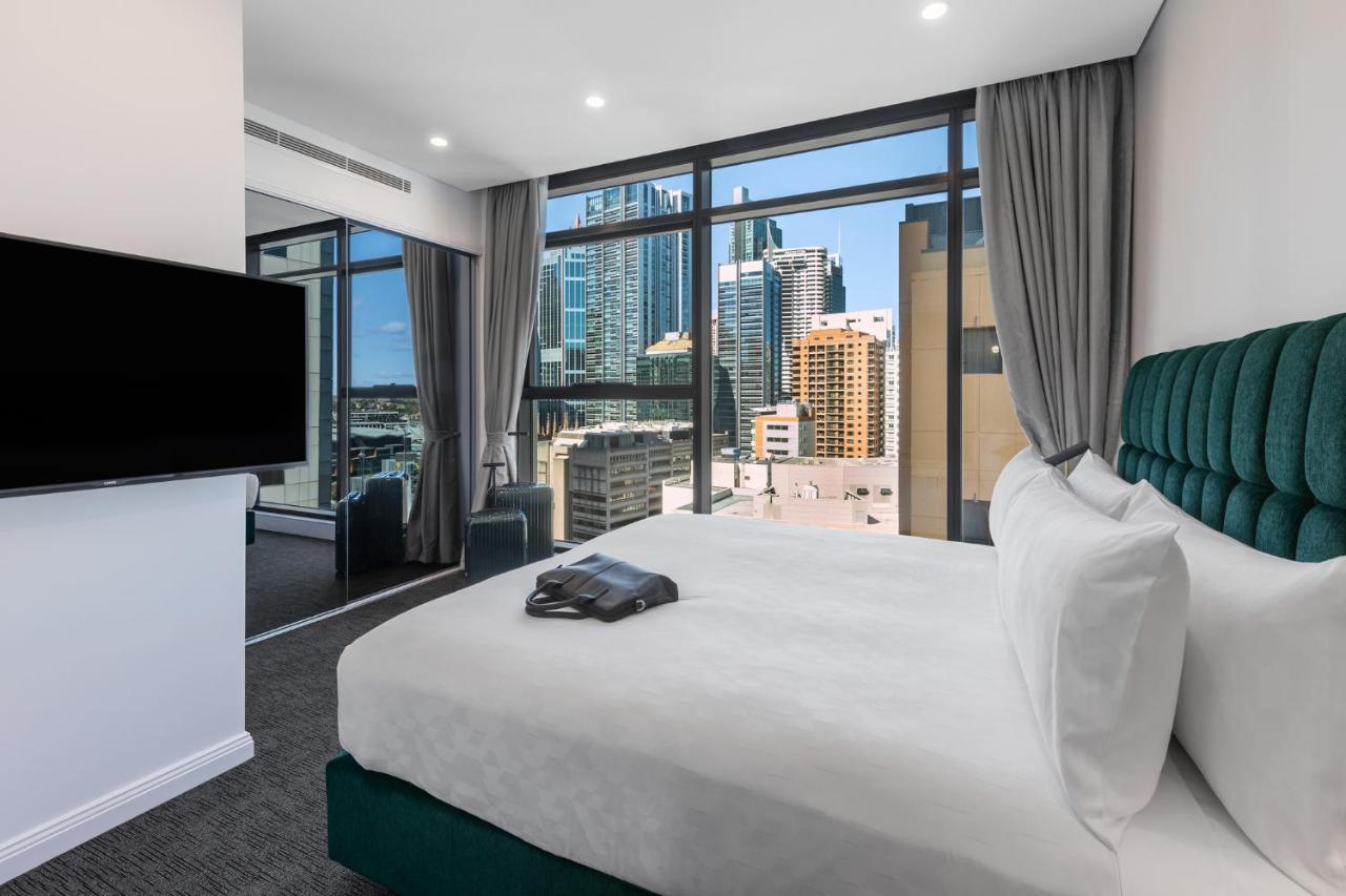 Meriton Suites Sussex Street, Sydney Zewnętrze zdjęcie The photo shows a modern hotel room featuring a large bed with white linens and a green upholstered headboard. There is a sleek television mounted on the wall opposite the bed, and the room has large windows that provide a view of a cityscape, includ