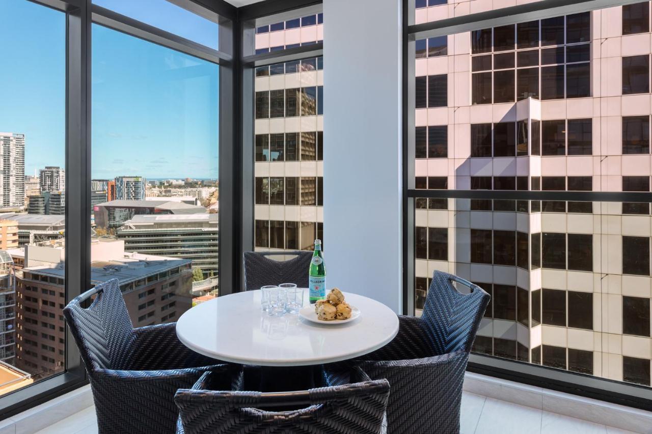 Meriton Suites Sussex Street, Sydney Zewnętrze zdjęcie The photo shows a modern dining space with a round white table set for four. There are four black woven chairs around the table. On the table, there's a bottle of sparkling water, two glasses, and a small plate with several cookies. The large windows