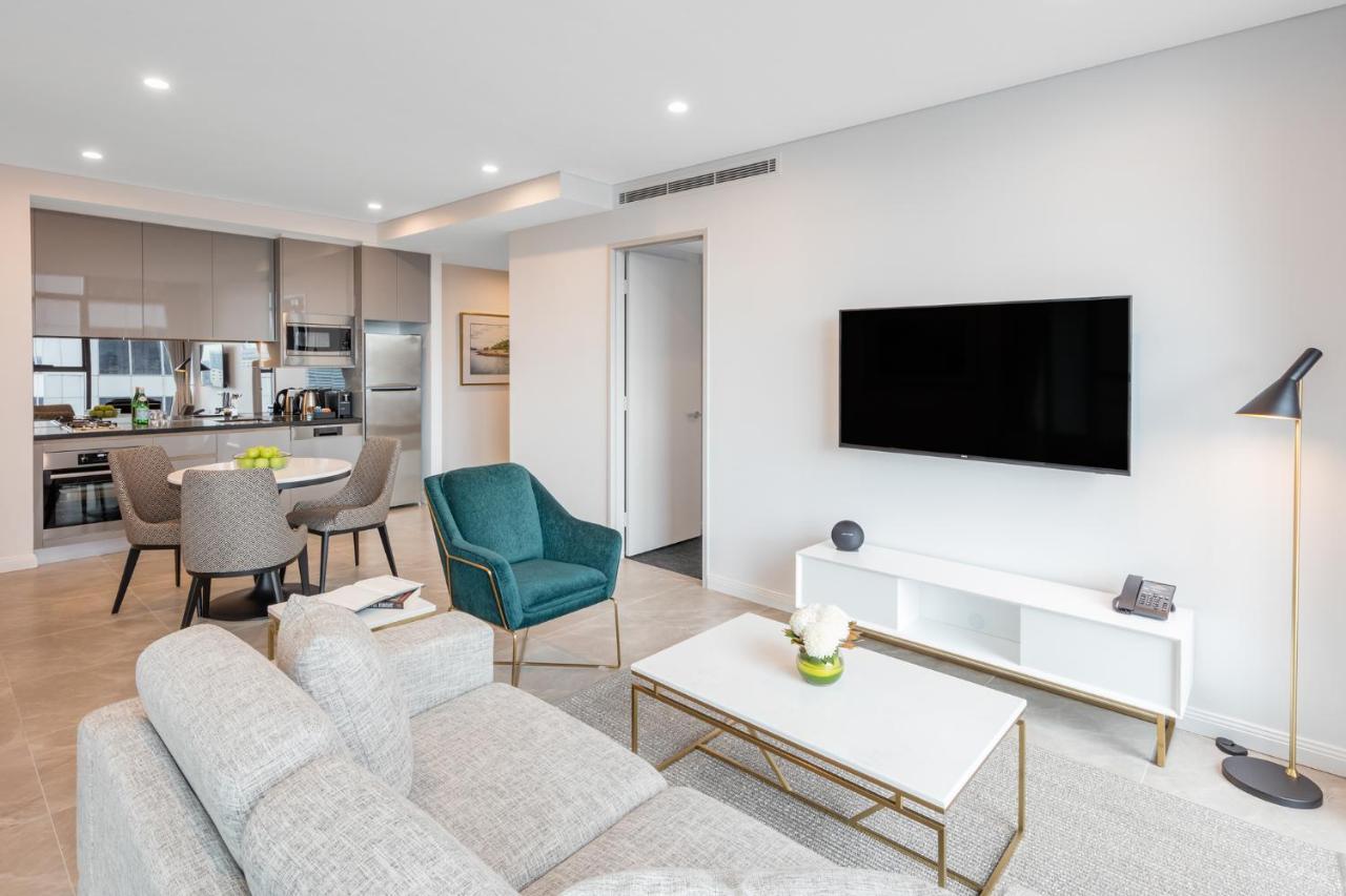 Meriton Suites Sussex Street, Sydney Zewnętrze zdjęcie The photo shows a modern living room area. It features a light gray sofa and a coffee table with a decorative plant. There are two chairs, one in teal and a round one, positioned around the coffee table. A flat-screen TV is mounted on the wall, and t