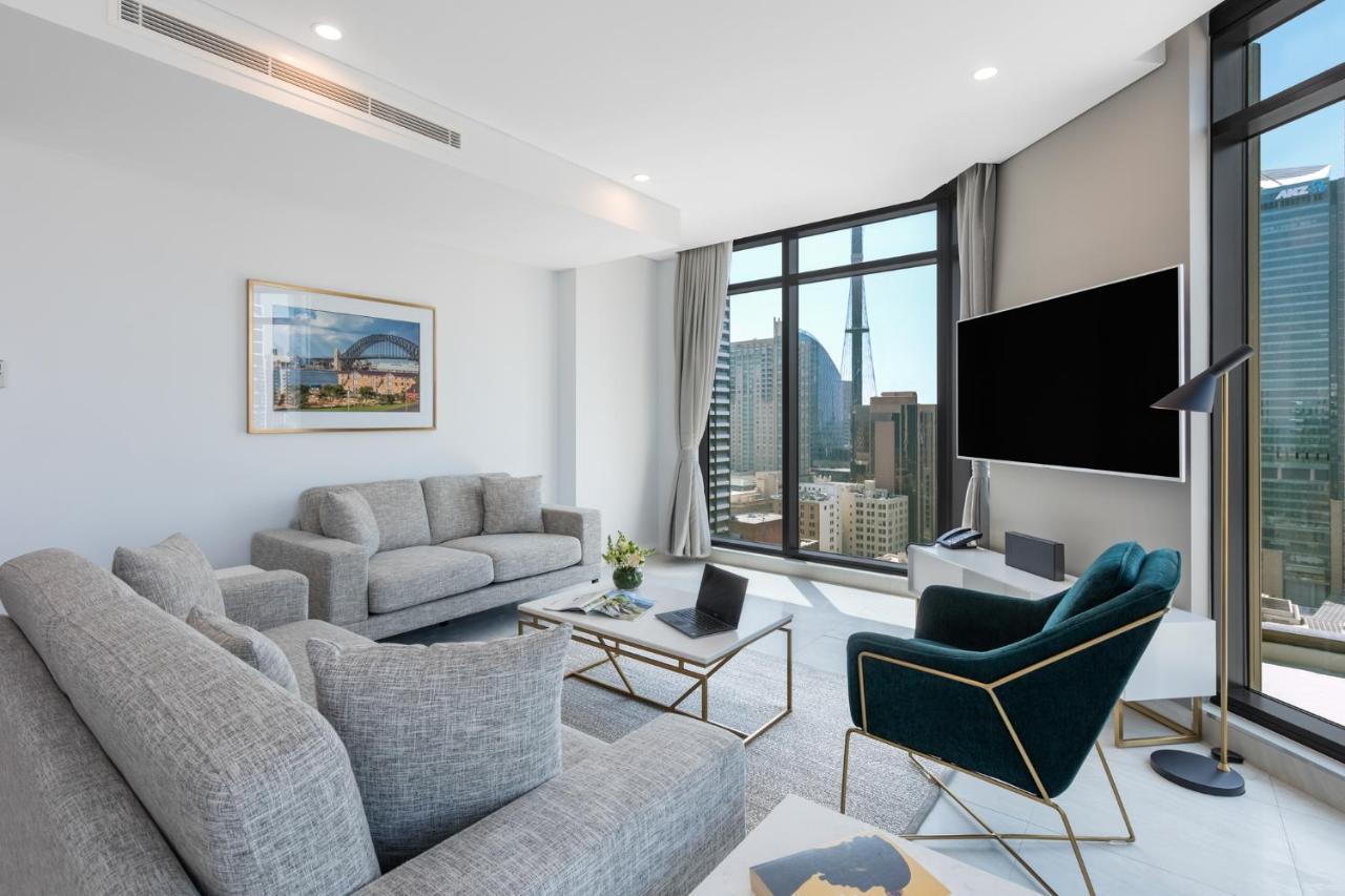 Meriton Suites Sussex Street, Sydney Zewnętrze zdjęcie The photo shows a modern living room with large windows offering a view of a city skyline. The room features two gray sofas and a stylish green accent chair. A sleek coffee table is placed in front of the sofas, alongside a laptop. There’s a flat-scr