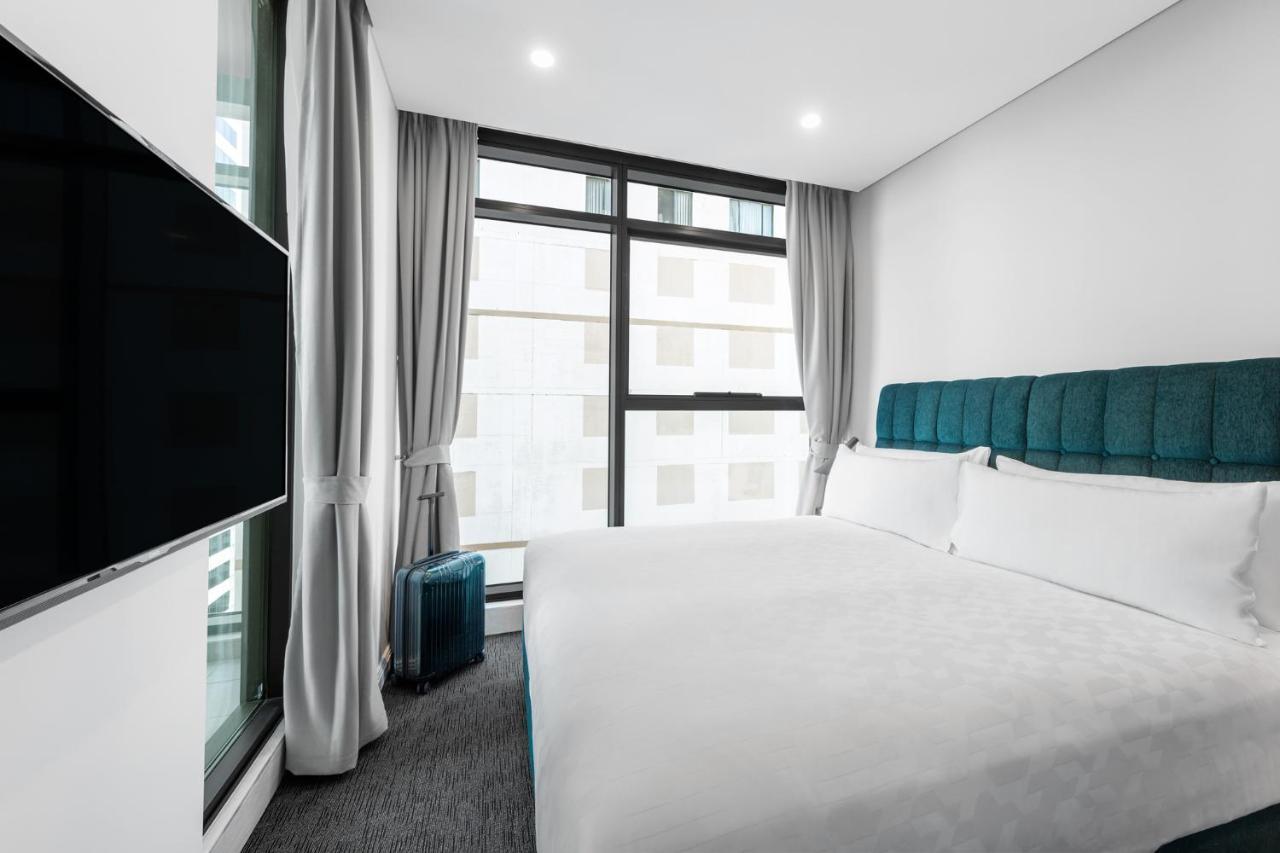 Meriton Suites Sussex Street, Sydney Zewnętrze zdjęcie The photo shows a modern hotel room featuring a large bed with a light-colored duvet and a blue upholstered headboard. There are two pillows on the bed. A large window allows natural light to enter the room, and gray curtains frame the window. Next t