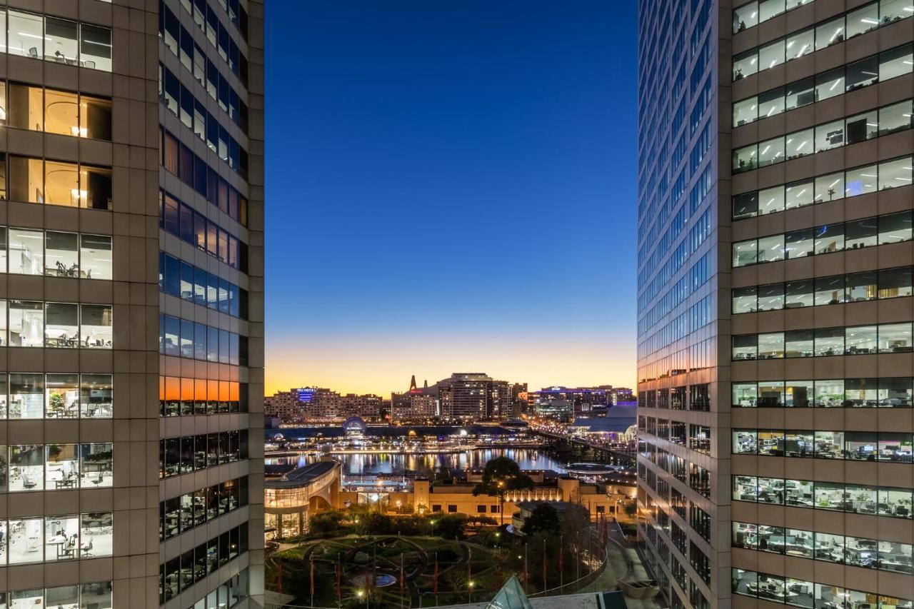 Meriton Suites Sussex Street, Sydney Zewnętrze zdjęcie The photo shows a view between two tall buildings, likely office towers, during twilight. On one side, there are glass windows reflecting the evening sky that transitions from blue to orange near the horizon. In the background, a cityscape is visible