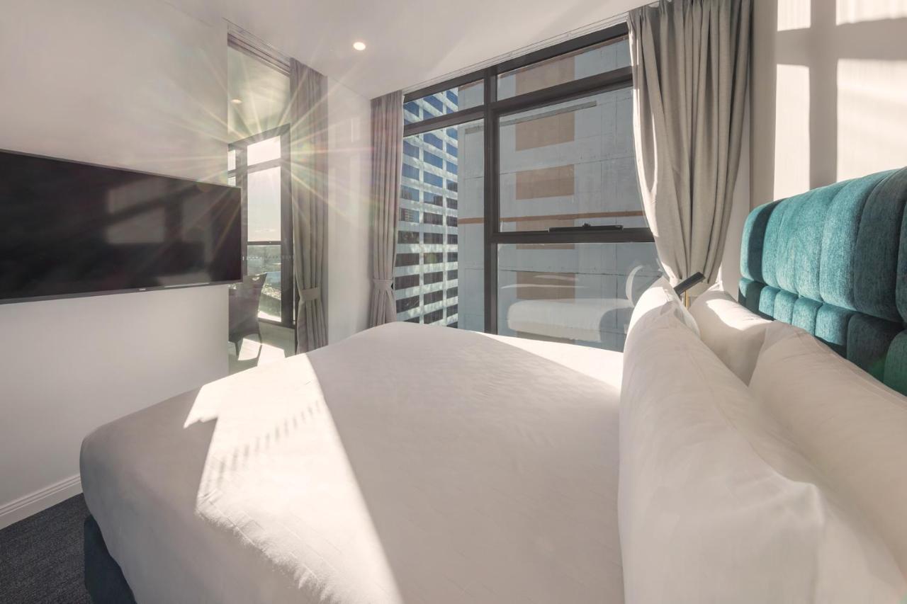 Meriton Suites Sussex Street, Sydney Zewnętrze zdjęcie The photo shows a modern hotel room with a neatly made bed featuring white bedding and a plush, tufted headboard in a shade of blue. Natural light is streaming through large windows, which are covered partially by light gray curtains. A flat-screen t