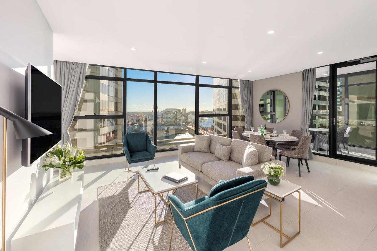 Meriton Suites Sussex Street, Sydney Zewnętrze zdjęcie The photo shows a modern and stylish living space. It features a spacious room with large windows that provide a bright view of the cityscape. The furniture includes a light gray sofa, two teal chairs, and a coffee table. There is a flat-screen TV mo