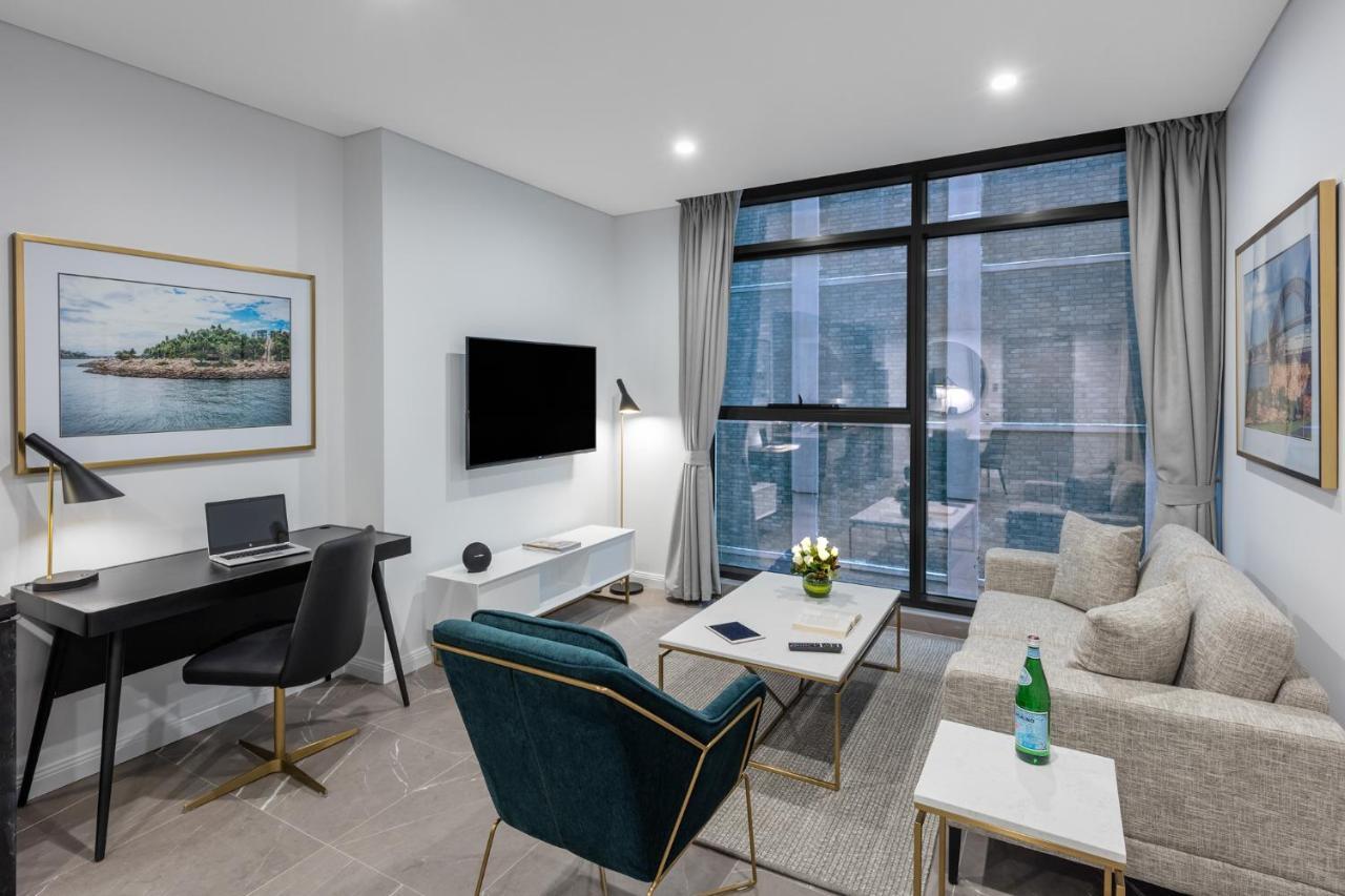Meriton Suites Sussex Street, Sydney Zewnętrze zdjęcie The photo shows a modern living area with a light color scheme. There is a comfortable seating arrangement that includes a beige sofa and an accent chair in teal. A coffee table is positioned in front of the sofa, with a small vase of flowers on it. 