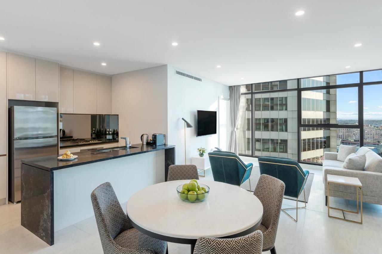 Meriton Suites Sussex Street, Sydney Zewnętrze zdjęcie The photo shows a modern and stylish interior space, likely a living room and kitchen area. The kitchen features sleek cabinetry and a countertop, with appliances visible, including a coffee maker. There is a round dining table with gray chairs, set 