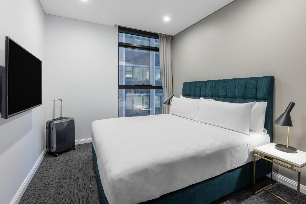 Meriton Suites Sussex Street, Sydney Zewnętrze zdjęcie The photo shows a modern hotel room with a clean and minimalist design. There is a large, neatly made bed with white bedding and a teal-colored headboard. On one side of the bed, there is a small nightstand with a lamp. A sleek television is mounted 