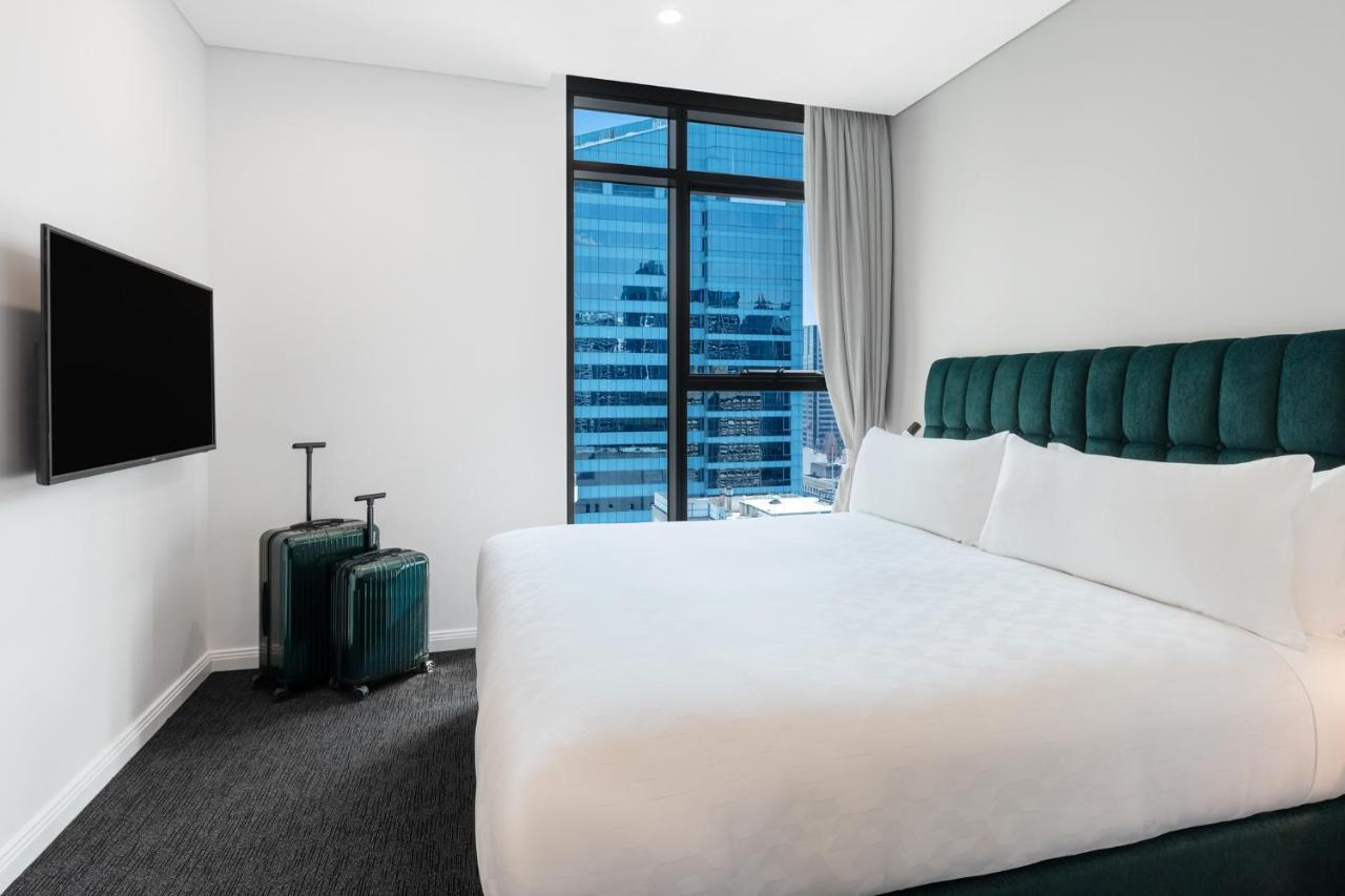Meriton Suites Sussex Street, Sydney Zewnętrze zdjęcie The photo shows a modern hotel room featuring a large bed with white bedding and a green upholstered headboard. There are two green suitcases placed beside the bed. A flat-screen TV is mounted on the wall opposite the bed. The room has a large window