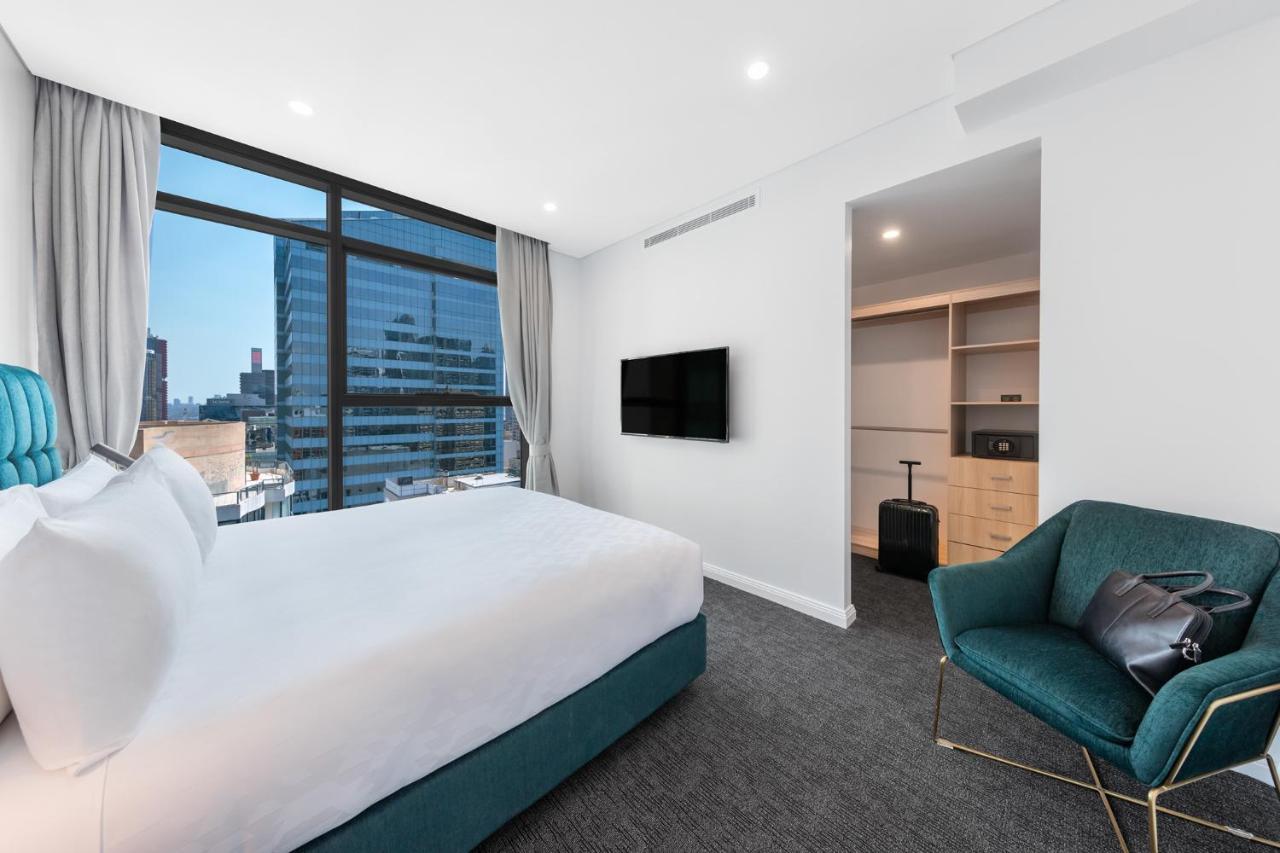 Meriton Suites Sussex Street, Sydney Zewnętrze zdjęcie The photo shows a modern hotel room. It features a large window that allows natural light to fill the space, offering a view of the city skyline. The room includes a neatly made bed with a white duvet and a teal-colored base. There is a small green a