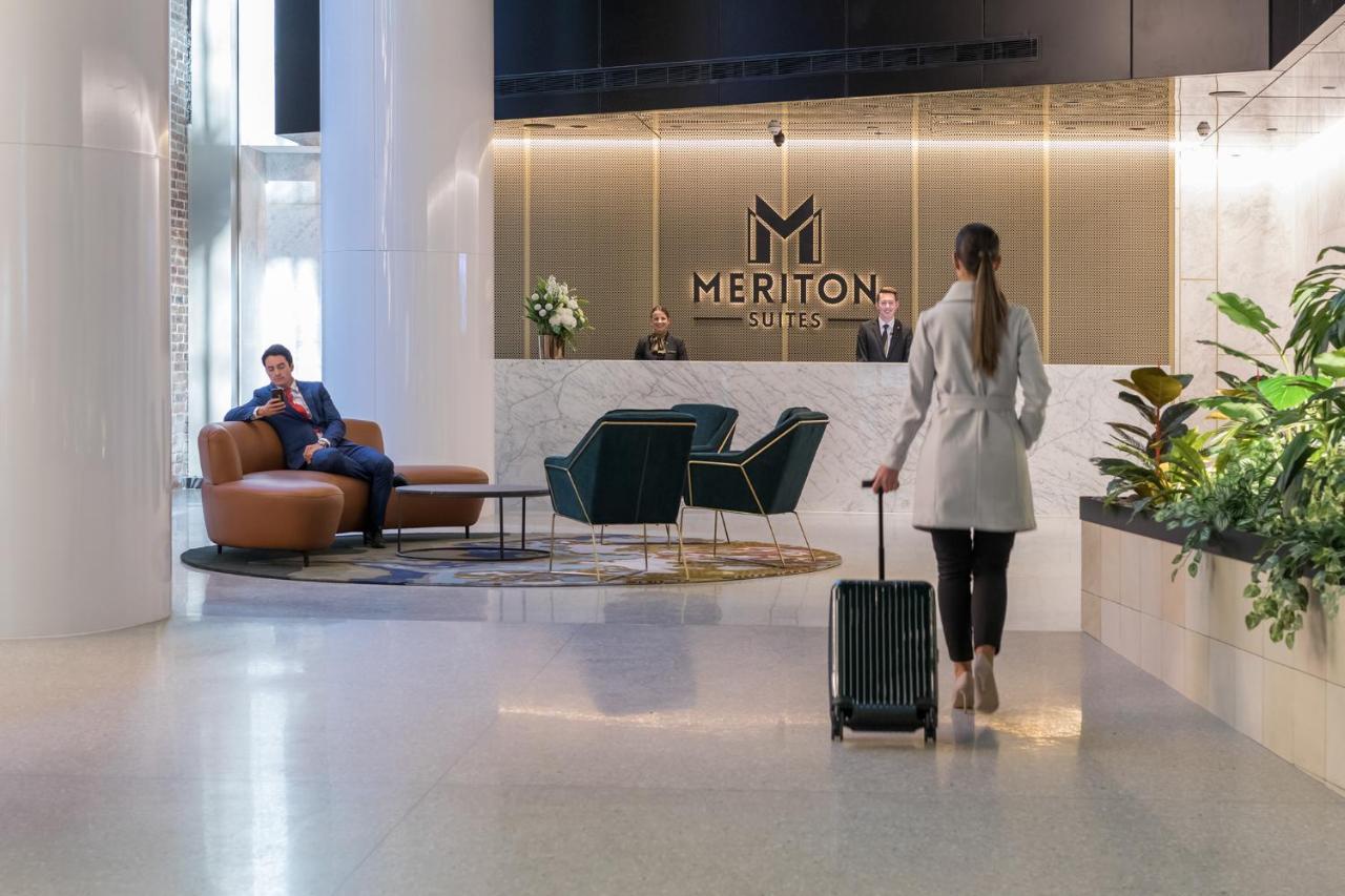 Meriton Suites Sussex Street, Sydney Zewnętrze zdjęcie The photo depicts a modern hotel lobby. In the foreground, a woman is walking towards the reception desk, pulling a suitcase behind her. She is dressed in a light-colored coat. To her left, there’s a comfortable seating area with a person sitting on 