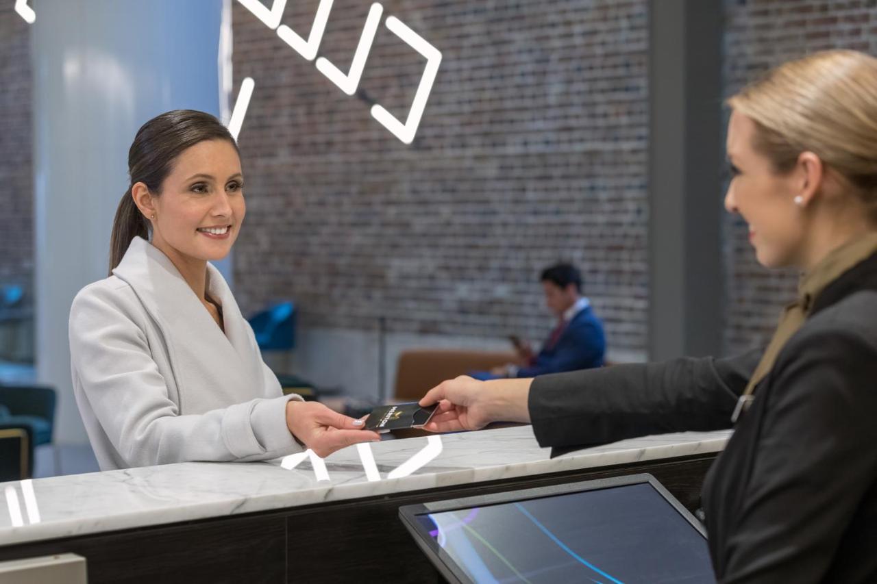 Meriton Suites Sussex Street, Sydney Zewnętrze zdjęcie The image depicts a scene at a hotel reception desk. A woman with long hair, dressed in a white outfit, is smiling as she hands her card to a receptionist, who is in a black uniform. The reception area features a modern design, and there is a person 