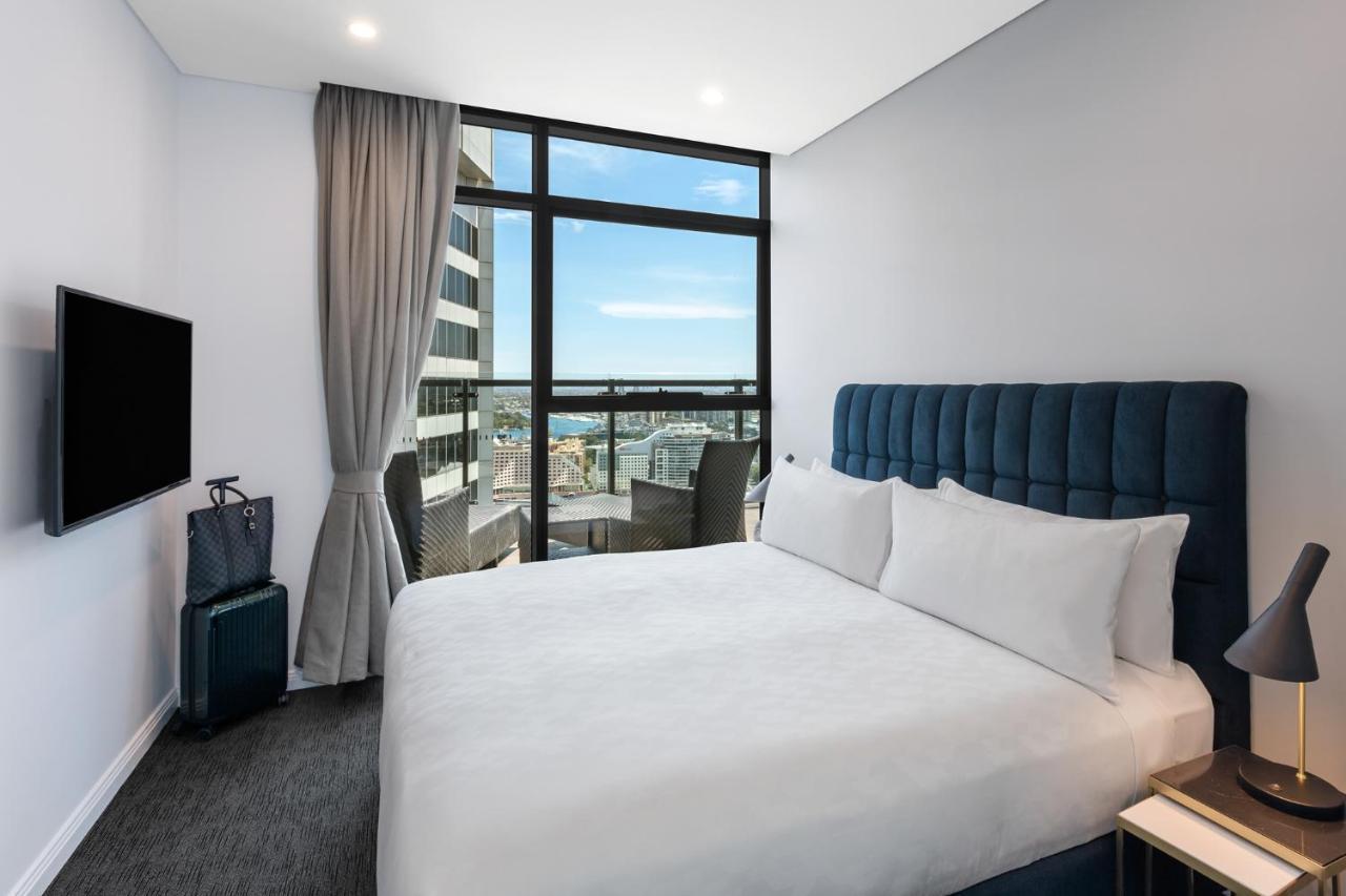 Meriton Suites Sussex Street, Sydney Zewnętrze zdjęcie The photo shows a modern hotel room featuring a double bed with white linens and a tufted blue headboard. There is a small bedside table next to the bed. A large window allows natural light to fill the room, offering a view of the city outside, inclu