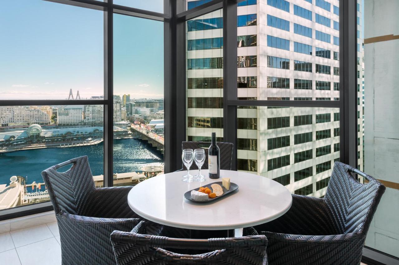 Meriton Suites Sussex Street, Sydney Zewnętrze zdjęcie The photo shows a modern dining area with a round table and four woven chairs. On the table, there are two wine glasses, a bottle of wine, and a plate with food items. The large windows provide a view of a cityscape featuring tall buildings and a bod