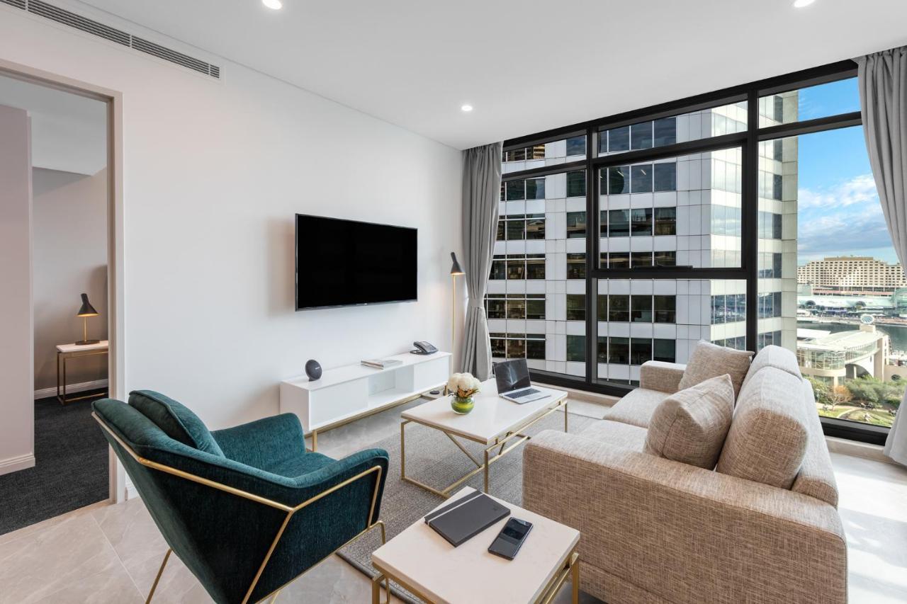 Meriton Suites Sussex Street, Sydney Zewnętrze zdjęcie The photo shows a modern living room with large windows that let in natural light, providing a view of a cityscape outside. The interior features a comfortable light-colored sofa and a stylish dark green armchair. A sleek TV is mounted on the wall op
