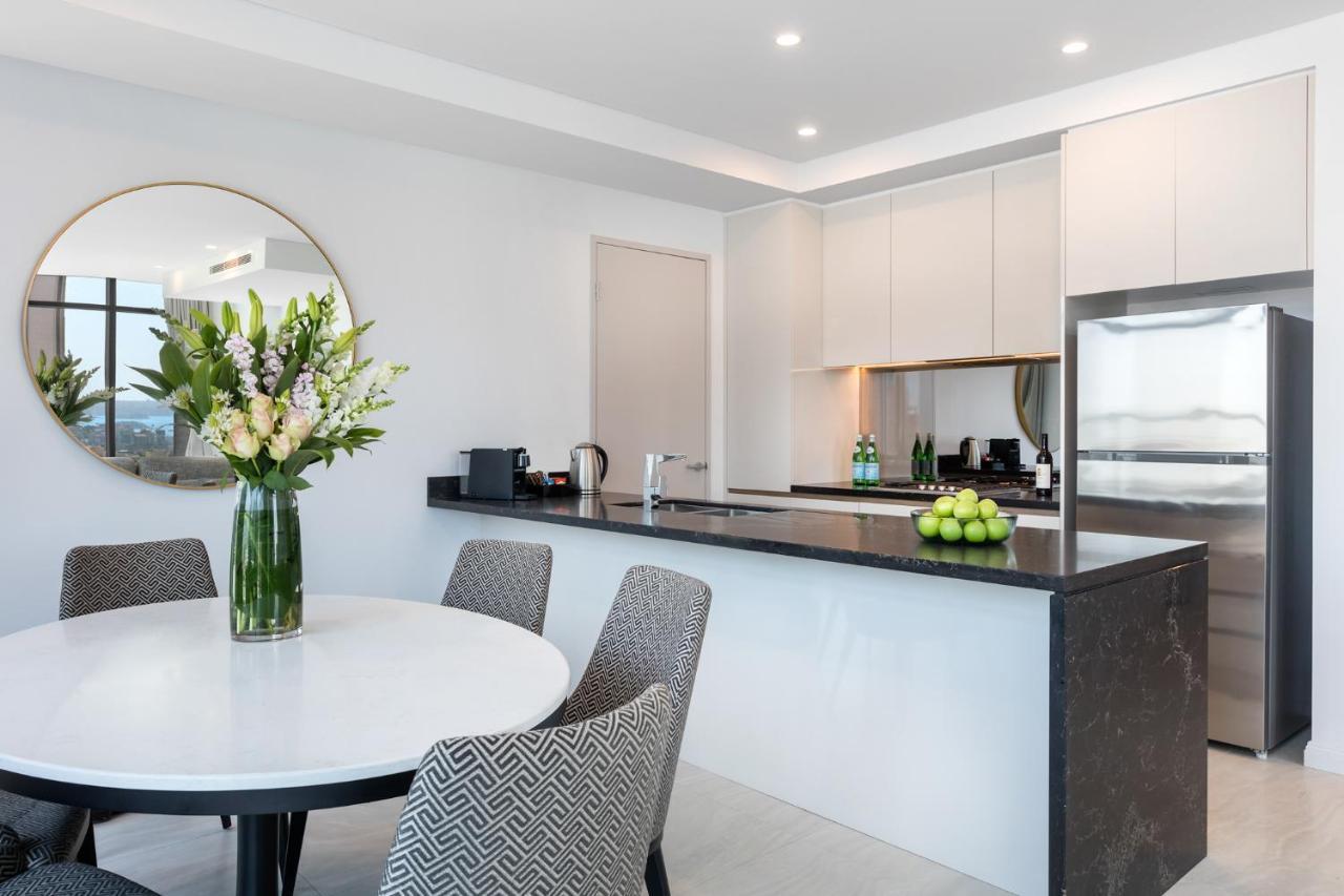 Meriton Suites Sussex Street, Sydney Zewnętrze zdjęcie The photo shows a modern kitchen and dining area. In the foreground, there's a circular dining table surrounded by four stylish chairs. A large vase filled with fresh flowers is placed on the table, adding a touch of color and elegance. 

In the kitc
