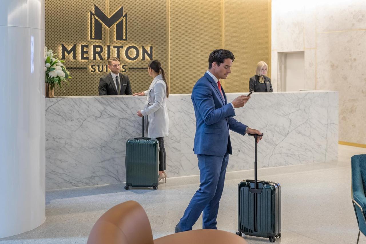 Meriton Suites Sussex Street, Sydney Zewnętrze zdjęcie The photo shows the interior of a hotel reception area, specifically at Meriton Suites. In the foreground, a man in a blue suit is standing with a suitcase, looking at his phone. Behind him, there are two staff members at the reception desk, one of w