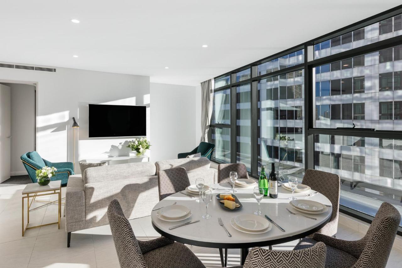 Meriton Suites Sussex Street, Sydney Zewnętrze zdjęcie The photo shows a modern and stylish living room and dining area. In the foreground, there is a round dining table set for a meal with plates, cutlery, and drinks. The seating arrangement includes comfortable chairs around the table. 

In the backgro