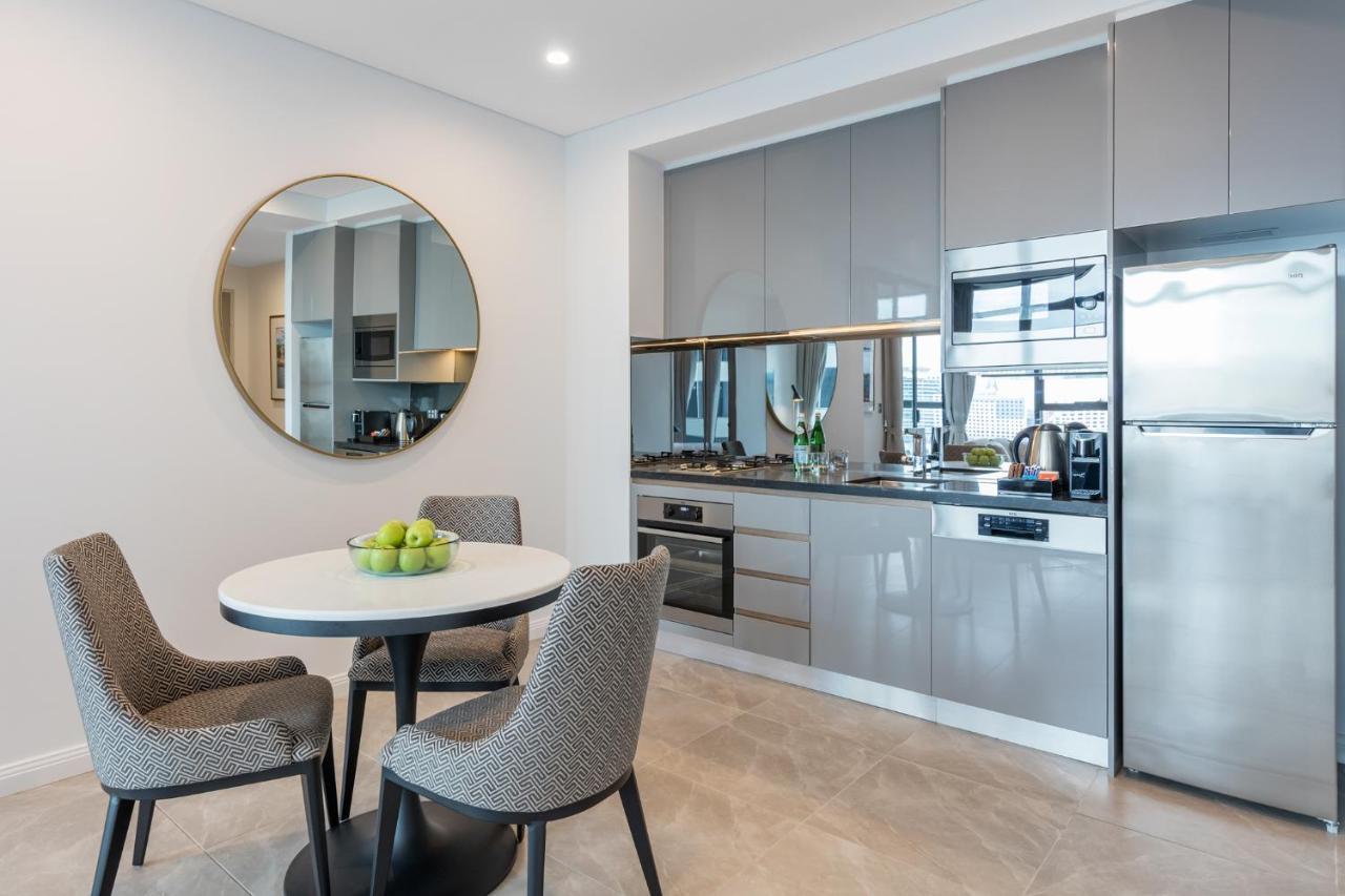 Meriton Suites Sussex Street, Sydney Zewnętrze zdjęcie The photo features a modern kitchen and dining area. The kitchen has sleek grey cabinets and countertops, with built-in appliances including an oven and possibly a cooktop. There's a large stainless steel refrigerator on the right. A round dining tab