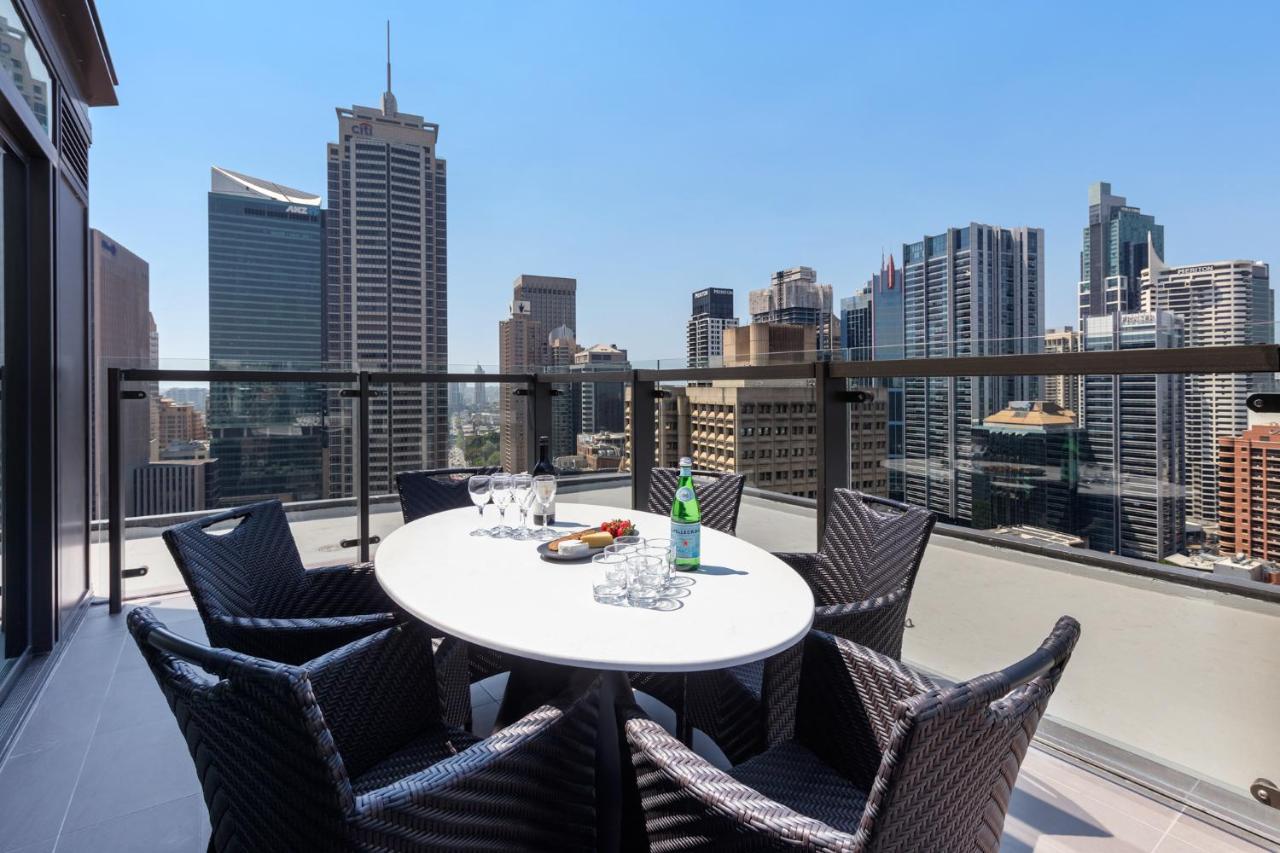 Meriton Suites Sussex Street, Sydney Zewnętrze zdjęcie The photo shows a stylish outdoor dining area on a balcony with a round white table surrounded by black wicker chairs. On the table, there are several glasses, a bottle of sparkling water, and a plate of snacks. In the background, there is a panorami
