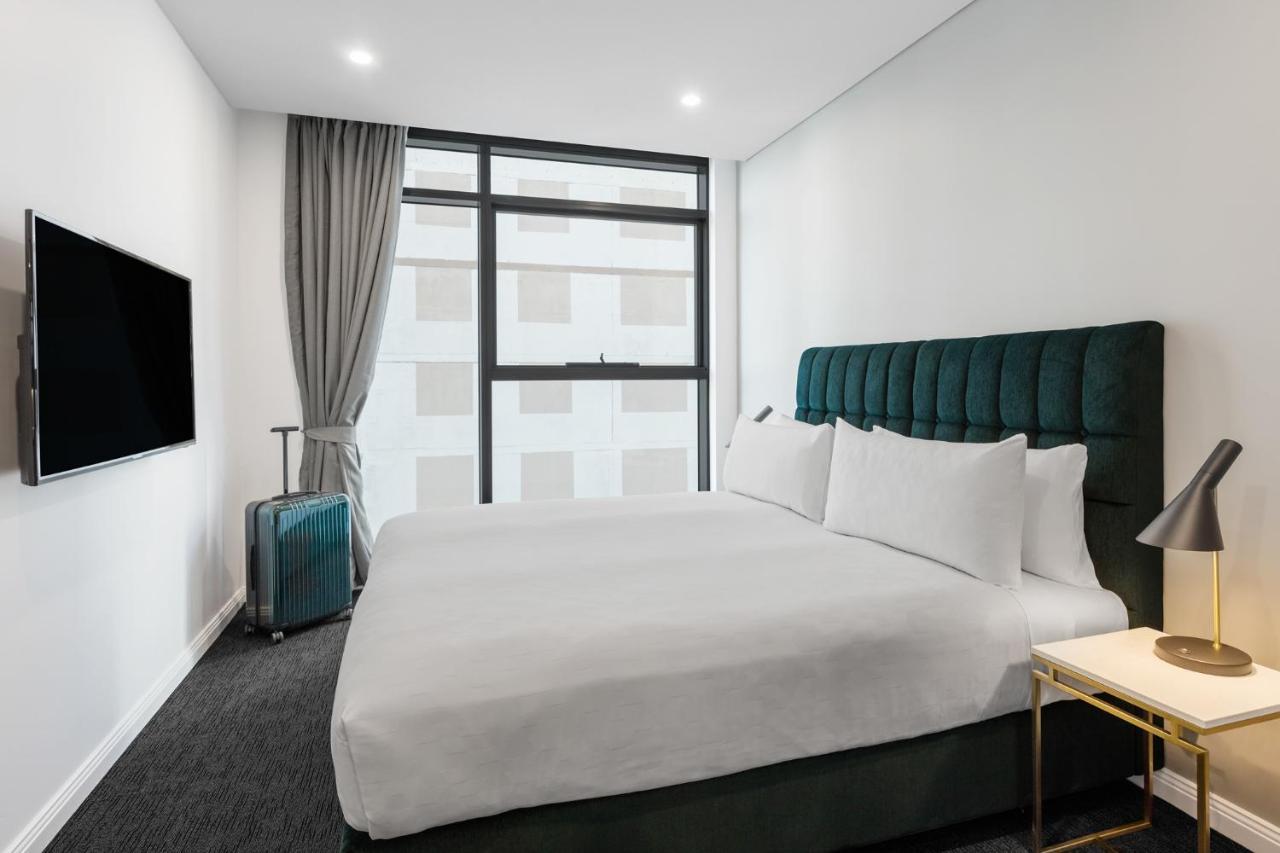 Meriton Suites Sussex Street, Sydney Zewnętrze zdjęcie The photo shows a modern hotel room. It features a large bed with a dark green tufted headboard and multiple white pillows. There's a nightstand with a lamp beside the bed. A sleek television is mounted on the wall opposite the bed, and a suitcase is