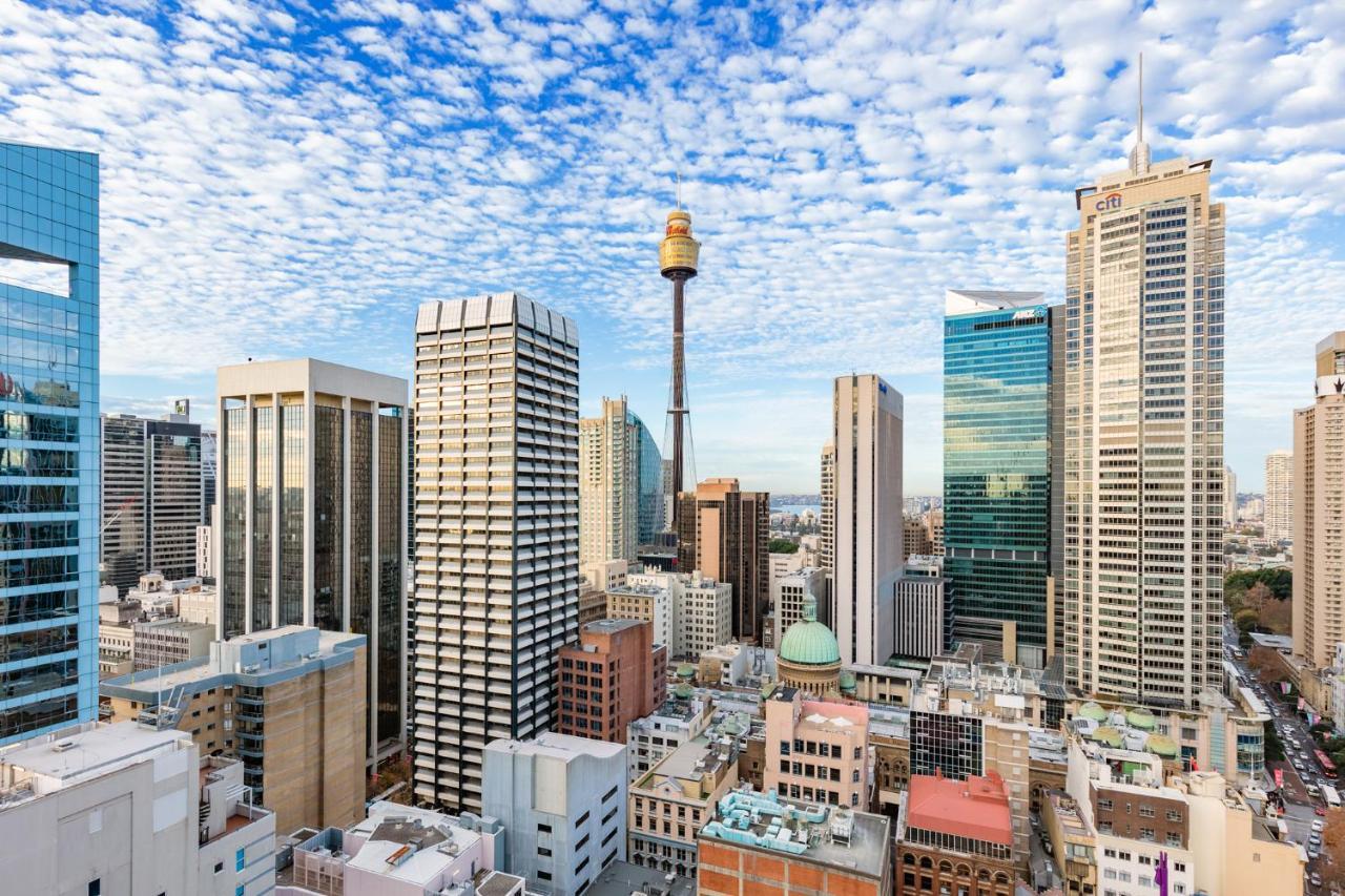 Meriton Suites Sussex Street, Sydney Zewnętrze zdjęcie The photo shows a panoramic view of a city skyline dominated by tall skyscrapers. In the center, there is a notable tower with a distinctive golden structure at its top, which is likely a landmark of the city. The buildings have a mix of modern glass
