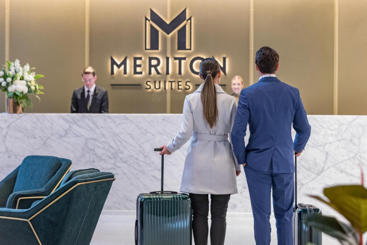 Meriton Suites Sussex Street, Sydney Zewnętrze zdjęcie The photo shows a couple standing at a reception area in a hotel named "Meriton Suites." They are both facing the reception desk, where staff members are visible. The man is dressed in a suit, and the woman is wearing a light-colored coat. They each 