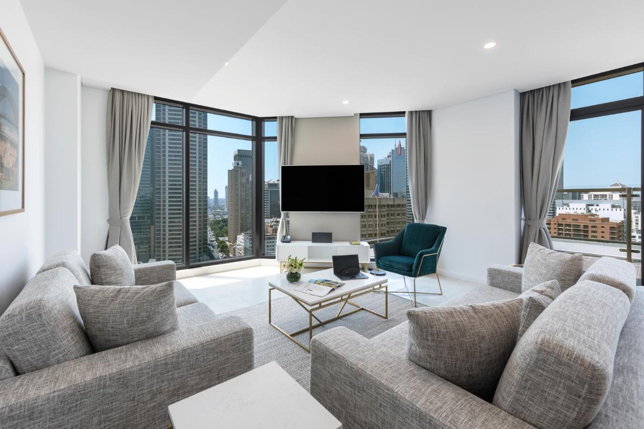 Meriton Suites Sussex Street, Sydney Zewnętrze zdjęcie The photo showcases a modern living room with large windows offering a view of a city skyline. The room features a light color palette with gray sofas arranged around a coffee table. A flat-screen TV is mounted on the wall, and there is a green armch