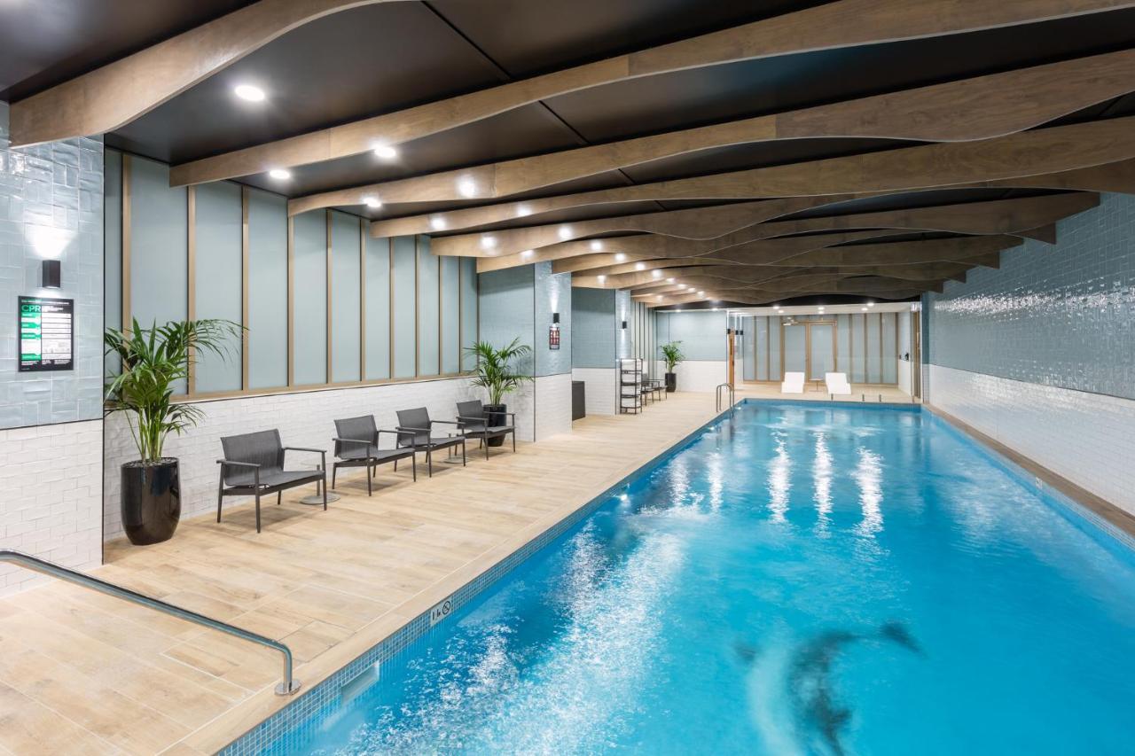 Meriton Suites Sussex Street, Sydney Zewnętrze zdjęcie The photo shows an indoor swimming pool area. The pool has clear blue water and is flanked by a wooden deck. There are several lounge chairs arranged along one side of the pool. The ceiling features a modern design with wooden beams and recessed ligh