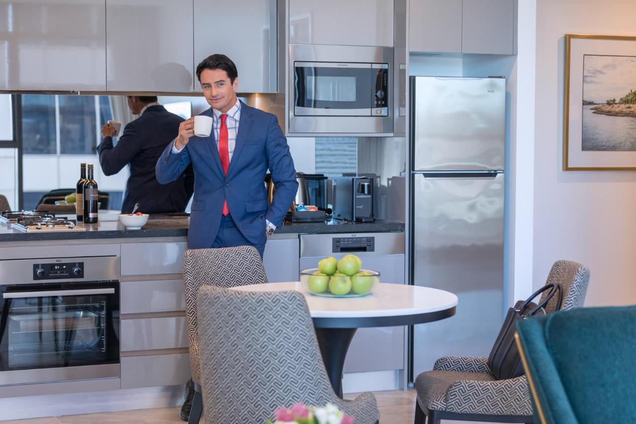 Meriton Suites Sussex Street, Sydney Zewnętrze zdjęcie The photo depicts a well-dressed man in a suit standing in a modern kitchen. He is holding a cup, possibly of coffee, and appears to be looking towards the camera. The kitchen features contemporary appliances, including a microwave and refrigerator. 
