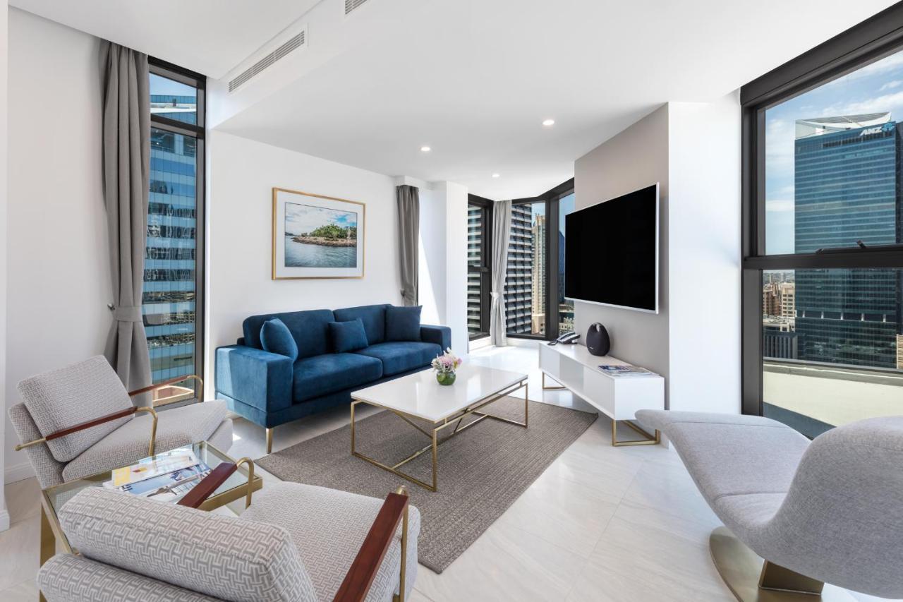 Meriton Suites Sussex Street, Sydney Zewnętrze zdjęcie The photo shows a modern living room featuring large windows that allow plenty of natural light. The room is elegantly designed with a blue sofa, a light-colored coffee table, and two armchairs. There’s a flat-screen TV mounted on the wall, and a pie